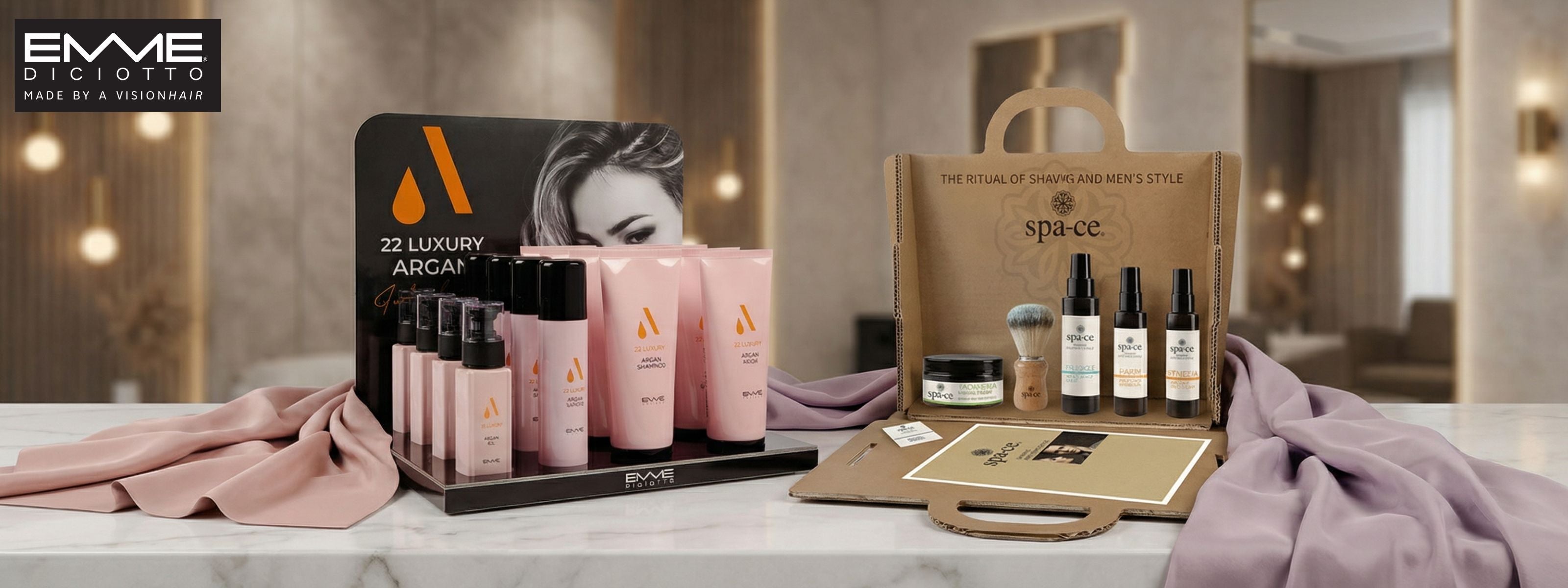 A luxurious display of Emme Diciotto pink argan oil shampoos, conditioners, and serums arranged on a draped silk cloth in a high-end salon. The scene includes professional silver round brushes and white branded boxes on a marble countertop.