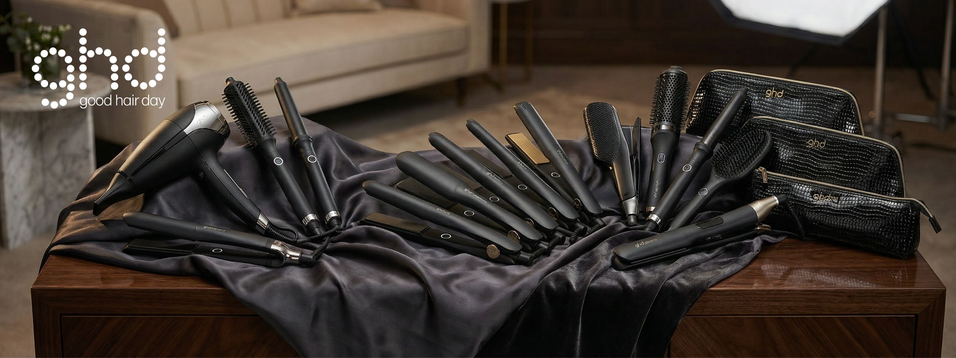 A wide array of black ghd professional hair styling tools, including blow dryers, flat irons, curling wands, and brushes, displayed on a dark silk cloth.